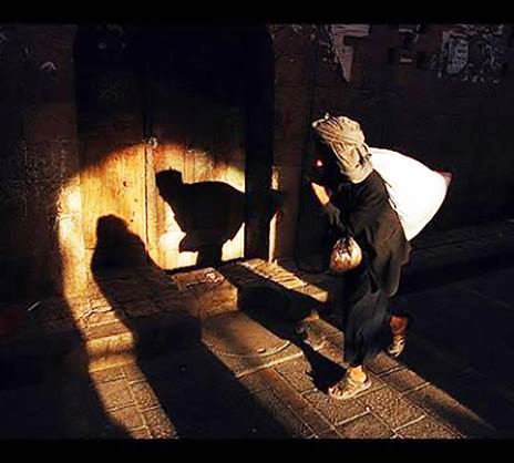 Una fotografía de un ladrón de espaldas cargando una gran bolsa blanca iluminado de repente por un foco potente que refleja su sombra en la parte inferior de una doble puerta de madera, toda esta escena contra un trasfondo negro de la noche. 