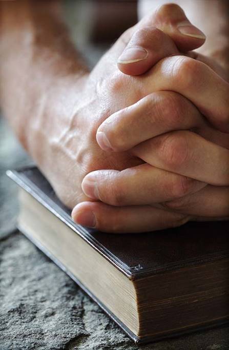 Clasped hands on a Bible resting on a solid rock in this photograph illustrate the subject We Invite You to Learn About the Church Christ Built, in iglesia-de-cristo.com.