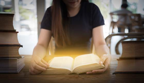 Imagen de una dama joven sentada a una mesa, con libros en cada lado de sus que sostienen una Biblia abierta de cuyas páginas emana una luz tenue blanca.