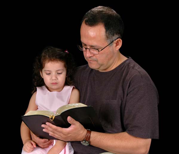 En esta fotografía un padre sostiene una Biblia abierta en su mano frente a su hijita sentada en su falda, ilustración para el tema Las iglesias de Cristo os saludan, en editoriallapaz.