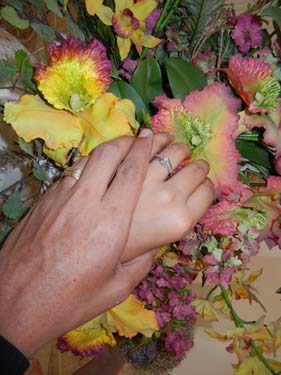 Esta fotografía de manos con anillos matrimoniales contra un trasfondo de flores ilustra la Ceremonia de boda de texto completo Dos radiantes luces se funden en una. Tres preguntas claves, en editoriallapaz.