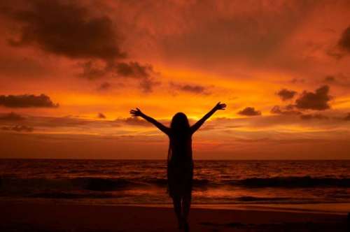 Esta fotografía de una dama con los brazos levantados en gesto de admiración y adoración, parada en una playa y contemplando un atardecer espectacular, ilustra los himnos a capela, a voces, disponibles en alabanzas.info.
