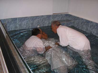 Una fotografía de dos varones vestidos de blanco en un bautisterio grande en el acto de sumergir a una persona, también vestida de blanco, en agua para el perdón de pecados.