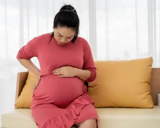 Una fotografía de una mujer latina joven encinta sentada sobre un sofá con una almohada amarilla frente a cortinas blancas, contemplando ella su barriga grande.