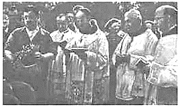 Fotografía en blanco y negro de varios clérigos católicos con vestimentas litúrgicas durante una ceremonia religiosa, uno de ellos leyendo un libro.
