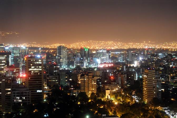 Fotografía panorámica de la enorme Ciudad de México de noche, cuyas tantísimas luces artificiales crean un reflejor en los cielos que la luna y las estrellas apenas se ven.