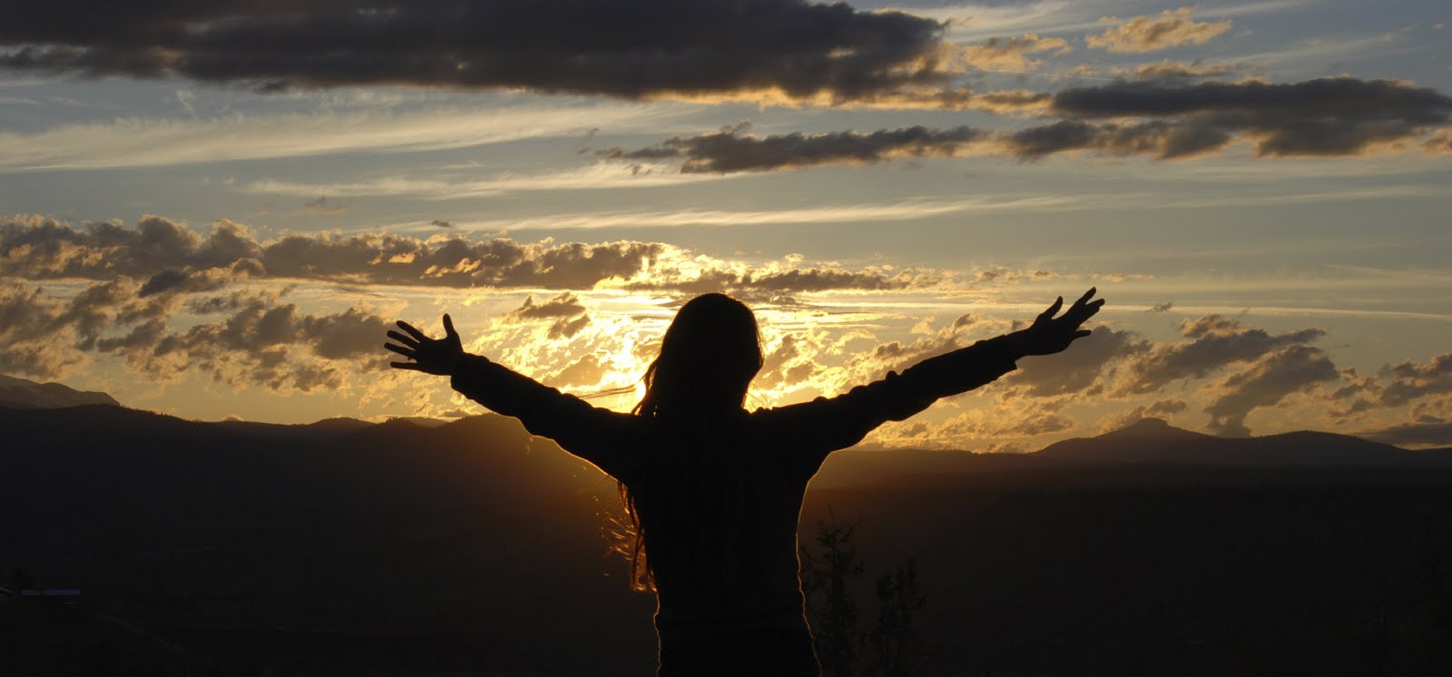 Fotografía de una dama de espaldas con sus brazos extendidos frente al sol que se pone sobre montañas ilustra el tema Ateos en el Edén en editoriallapaz.org.