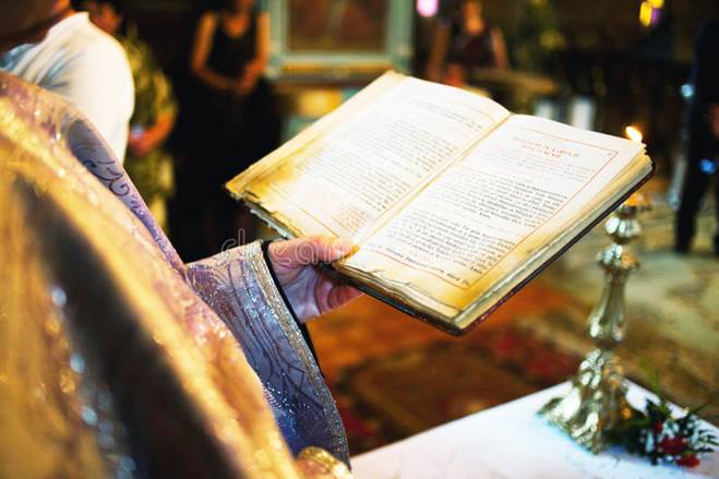 Fotografía de una Biblia grande abierta ante un sacerdote católico que viste lujosas vestimentas, con artefactos de plata, etcétera, en una mesa.