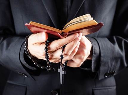 Fotografía de las manos de un sacerdote católico que sostiene en sus manos un rosario con un crucifijo y una Biblia abierta.