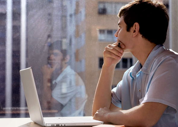 Esta fotografía de un varón joven sentado frente a una computadora y mirando, meditabundo, hacia la derecha su reflexión en ventanas de cristal, ilustra el intercambio con un chileno profesional que critica a las iglesias, llamando a la tolerancia religiosa