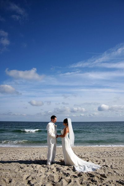 Esta fotograf'ia de una pareja vestida de boda parada en una playa bajo un cielo azul ilustra la Lista de bodas en editoriallapaz.org.