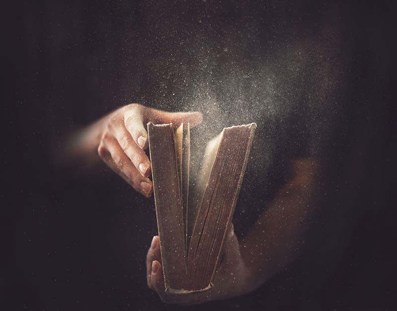 En esta fotografía matizada por ciertos elementos de misterio y espiritualidad, dos manos masculinas sostienen una antigua Biblia grande entreabierta, visibles solo las manos, la Biblia y una luz tenue blanca que ilumina partículas de polvo, todo contra un trasfondo de tonalidades de marón y negro, ilustración para el tema Falsificando al firma de Dios, en EditorialLaPaz.org.