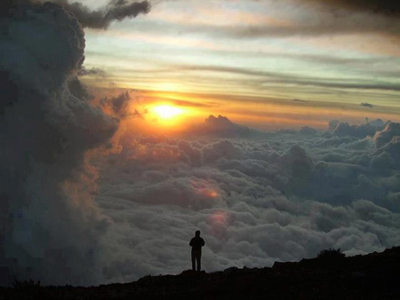 Un varón solitario, parado en la ladera de una montaña alta, y contemplando delante de él una vasta expansión de nubes y cielos, con el sol en el horizonte, ilustra el mensaje en audio Soñando despierto, por Alfonso Estrella, de la Iglesia de Cristo en Bayamón, Puerto Rico.
