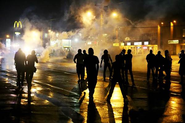 Una fotografía de doce figuras humanas en silueta que observan la quema de vehículos y tiendas en protestas sociales-raciales, para el tema Justicia después de la muerte.