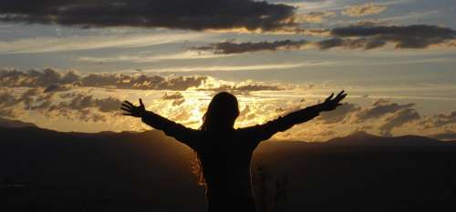 Fotografía de una dama de espaldas con sus brazos extendidos frente al sol que se pone sobre montañas ilustra el tema Ateos en el Edén en editoriallapaz.org.