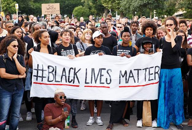 Una fotografía de una muchedumbre de personas que sostiene de ellas una pancarta grande con las palabreas en inglés Black Lives Matter, ilustración para el ensayo El color de vida humana.